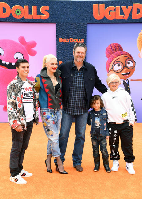 gwen and blake stand smiling on a bright orange carpet with three children standing with them and they all have an arm wrapped around each other and are dressed in casual clothing but gwen wears black stiletto heels