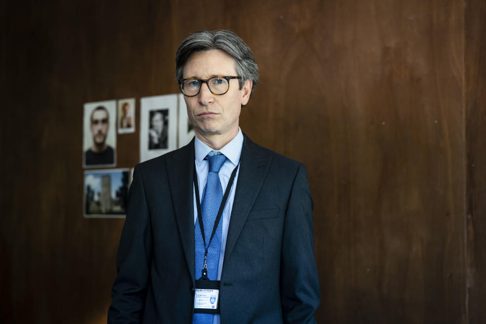 Jonathan Aris as DCI Mark Glover wearing glasses with pictures of Ben Field on the wall behind him..