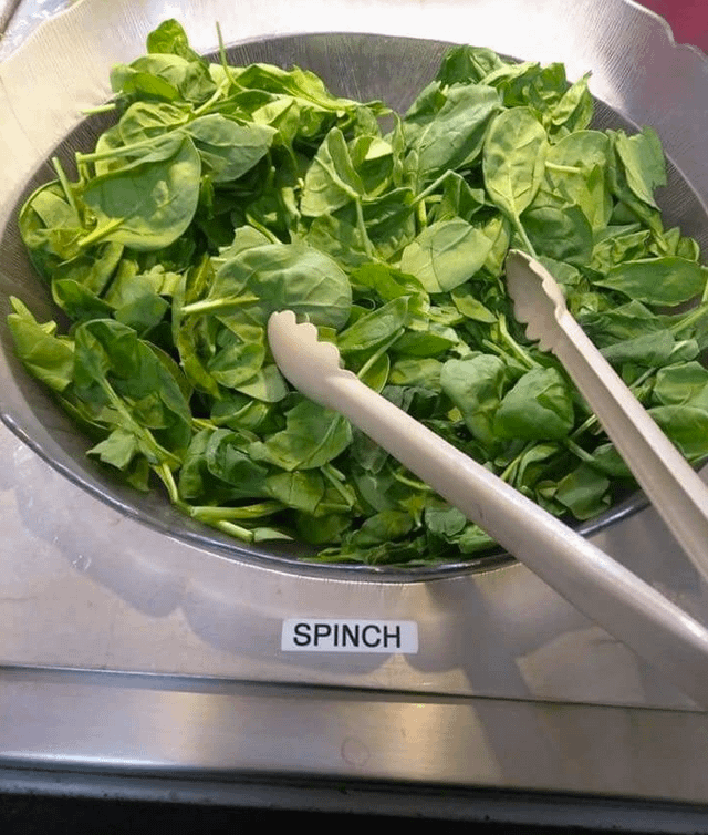 A bowl of fresh spinach leaves with a pair of tongs, and a label with a misspelled word "SPINCH" in front