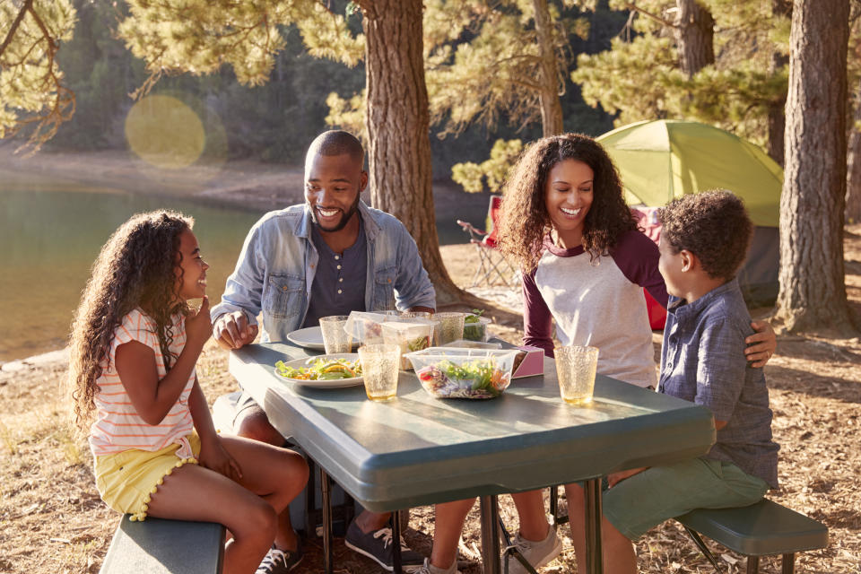 Man, woman, young girl, and young boy sitting at picnic table with food laid out on top of it.