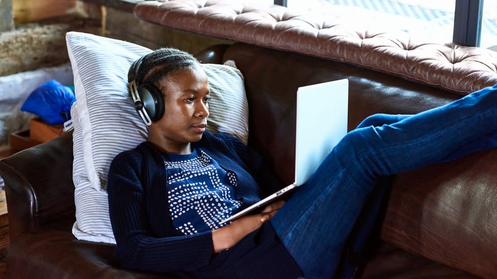 Una joven usa su laptop y lleva audífonos mientras está acostada en un sofá