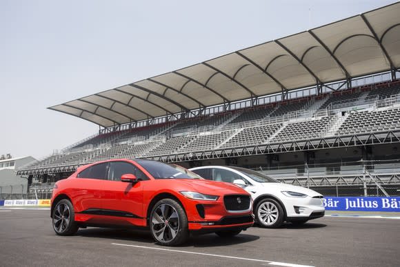 A red Jaguar I-Pace and a white Tesla Model X SUV, side-by-side on a race track.