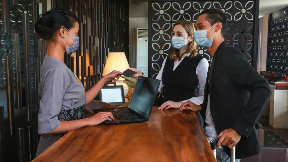 Couple and receptionist at counter in hotel wearing medical masks as precaution against virus.