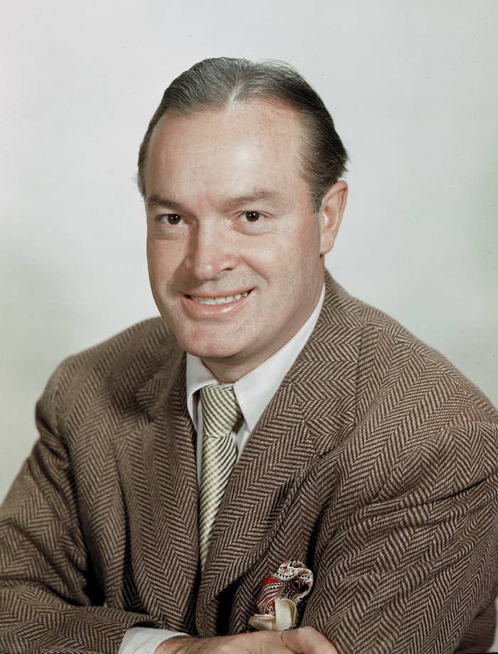 A man with slicked-back hair and a checkered jacket smiles at the camera