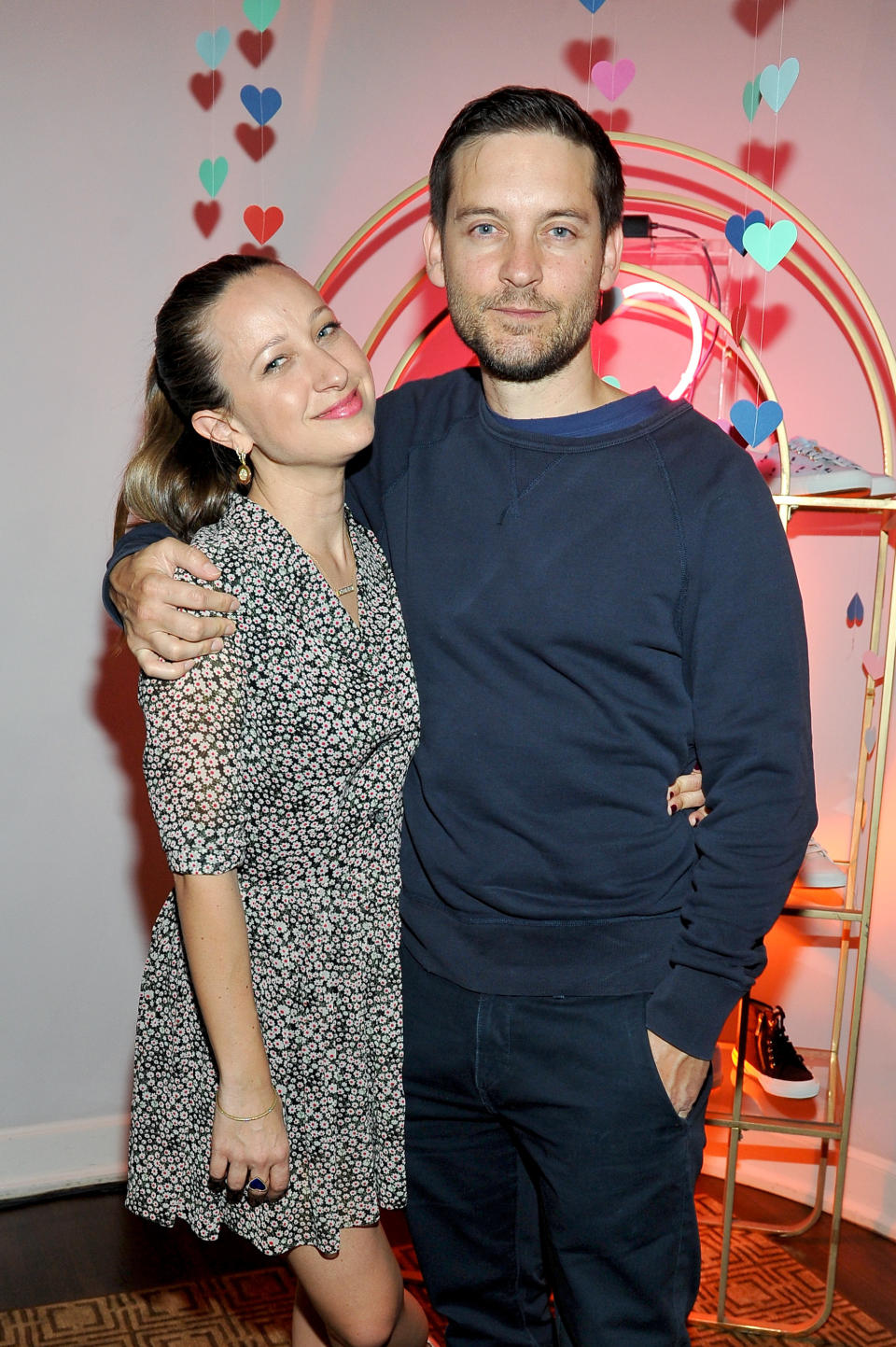 Jennifer Meyer and Tobey Maguire smiling and posing together, with Tobey's arm around Jennifer's shoulders. They are standing in front of a backdrop with heart decorations