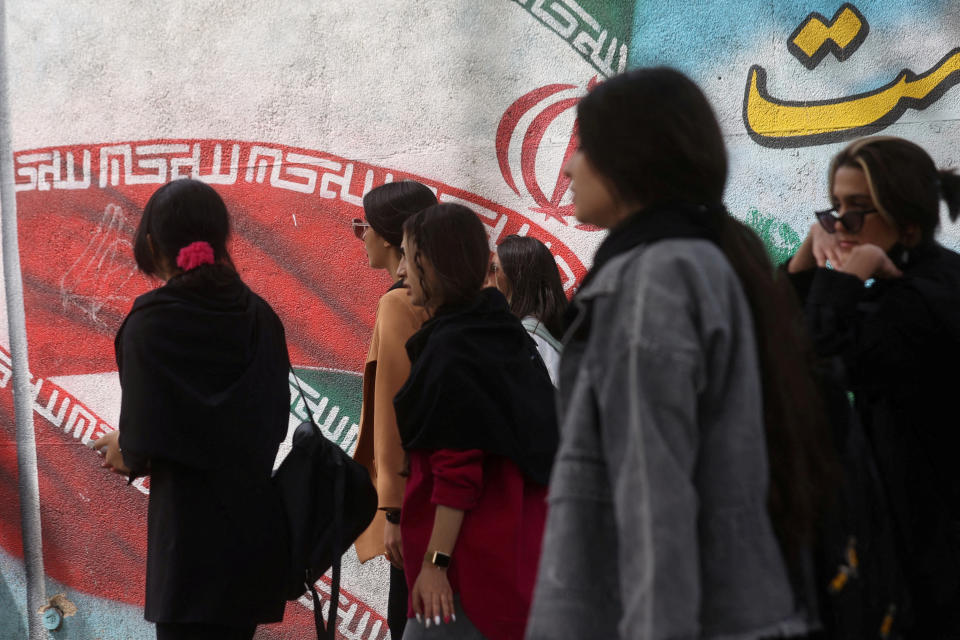 A group of Iranian women wearing Western dress and no chador veils, one with bare arms, pass a huge revolutionary mural. 