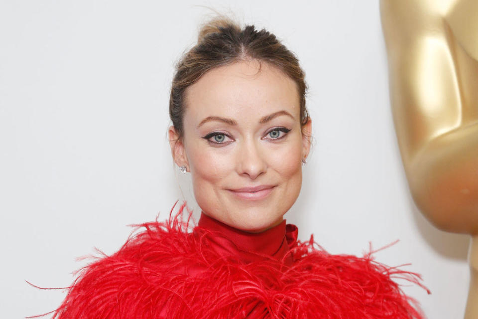 Olivia slightly smiling for cameras in a feathered outfit at an Oscars event