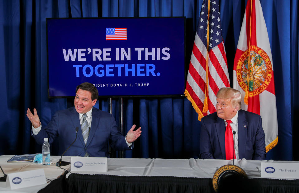 Ron DeSantis and Donald Trump sit at a table in front of microphones in front of flags and read on signs: We are in this together, President Donald J. Trump
