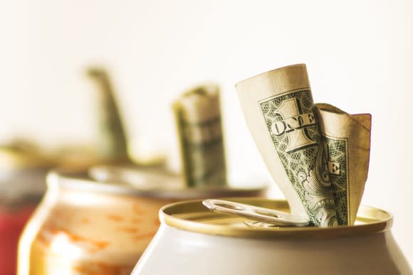 A close-up view of aluminum soda cans against a white background, with money placed inside the drinking spouts.