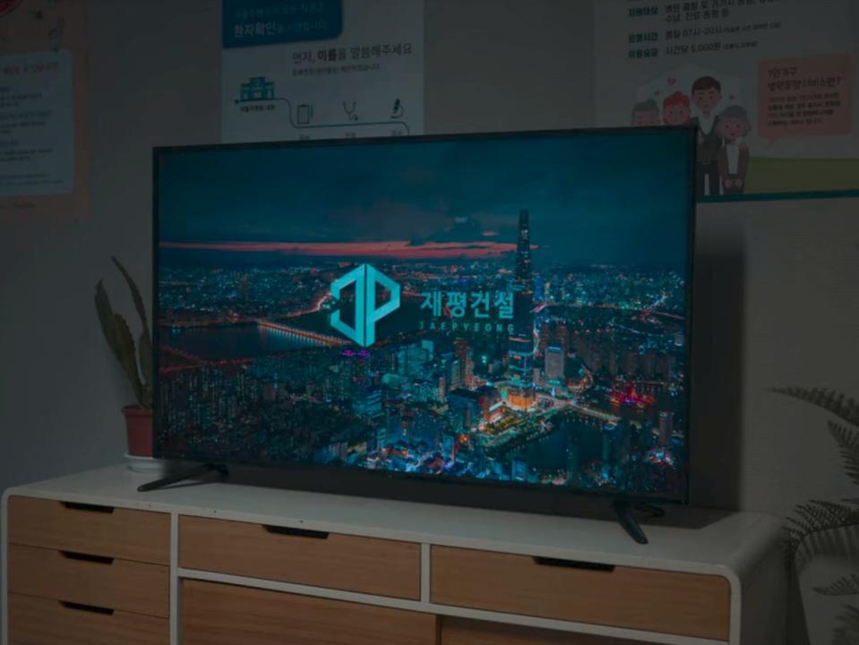 a television on a white tv stand, with hospital posters behind it with text in korean. the television is showing an advertisement with a shot of a cityscape and the logo for a company titled jaepyeong construction