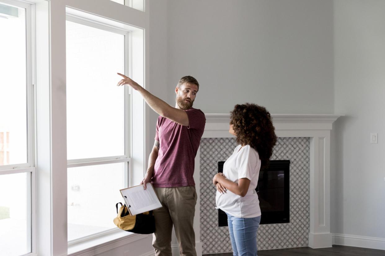 Serious home inspector or handyman points to something outside of a window while talking with pregnant homeowner.