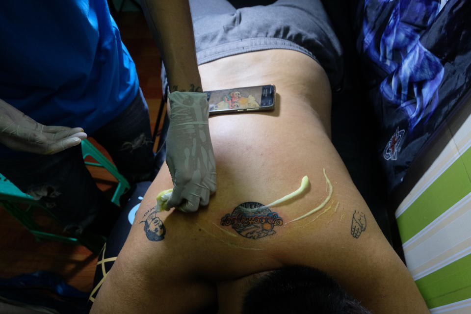 A tattoo artist wipes over a fresh tattoo with a design with Burmese text that reads "spring revolution" on the back of a man with other tattoos that depict the three-fingered protest salute and deposed Myanmar leader Aung San Suu Kyi in Yangon, Myanmar Thursday, March 25, 2021. (AP Photo)