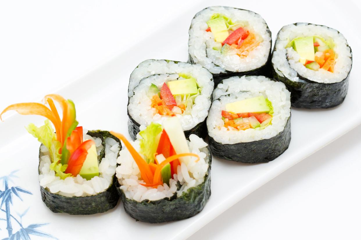 Six pieces of vegan sushi with avocado, carrots, lettuce and red pepper on a white porcelain rectangle plate with blue bamboo motif on the corner on a white background