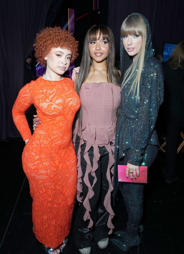 ice spice, pinkpantheress, and taylor swift stand together for a photo, ice spice wears an orange lace dress, pinkpantheress wears a dusty pink sleeveless top with dark pants, and taylor swift wears a black sparkly outfit with a hood