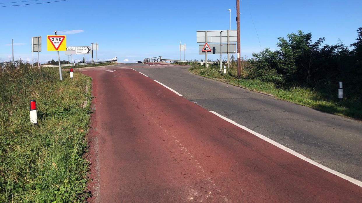 Boots Bridge approach in Manea, Cambridgeshire, looking up an incline on red-painted tarmac towards the bridge, with a "give way" warning sign 
 and another "quayside or river bank" sign showing a vehicle plunging in to water