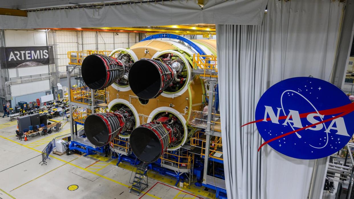  A rocket with four engines on top of a large construction structure. the logo for artemis is on the left (on a wall) and the meatball logo of nasa is on the right (on a curtain to the side of the rocket). 