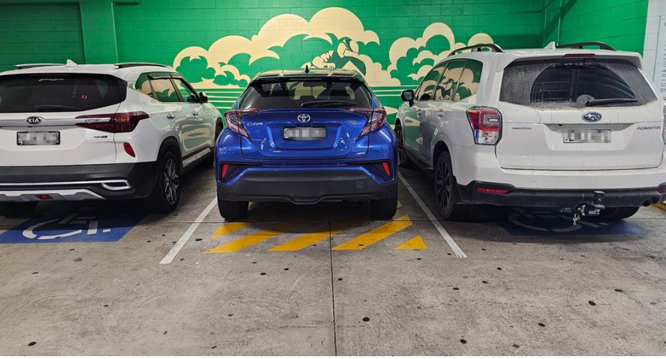 A car is seen here parked in between two disability bays, on top of yellow lines. 