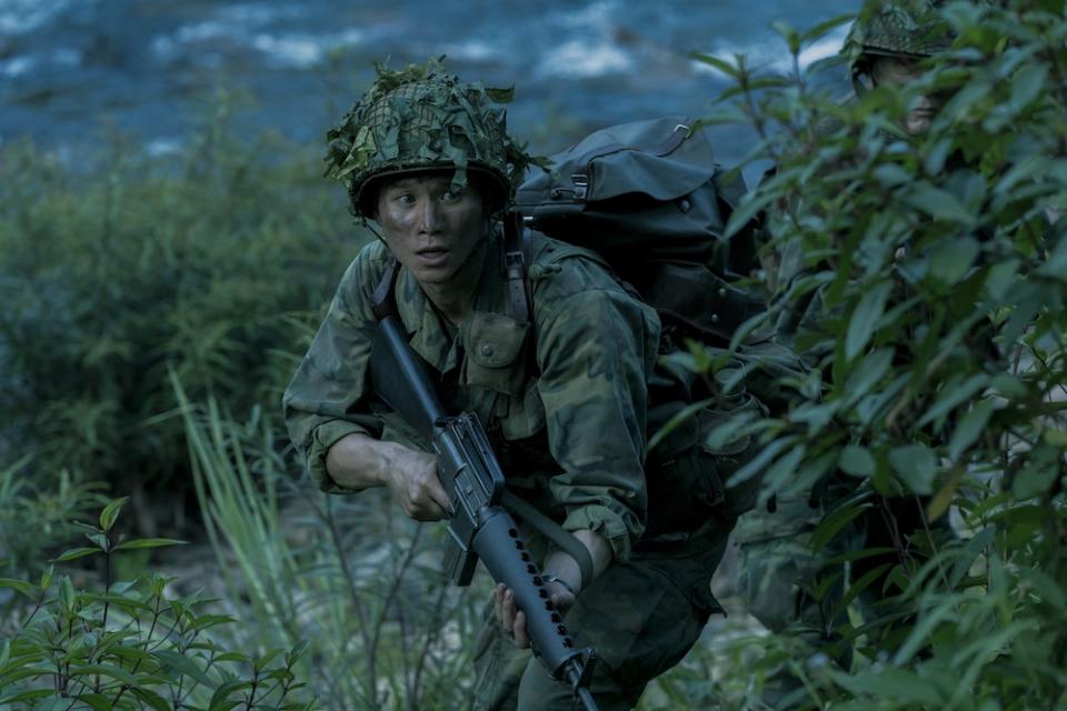 The Sympathizer stars Hoa Xuande as The Captain, shown here in green camoflauge carrying a rifle in the jungle