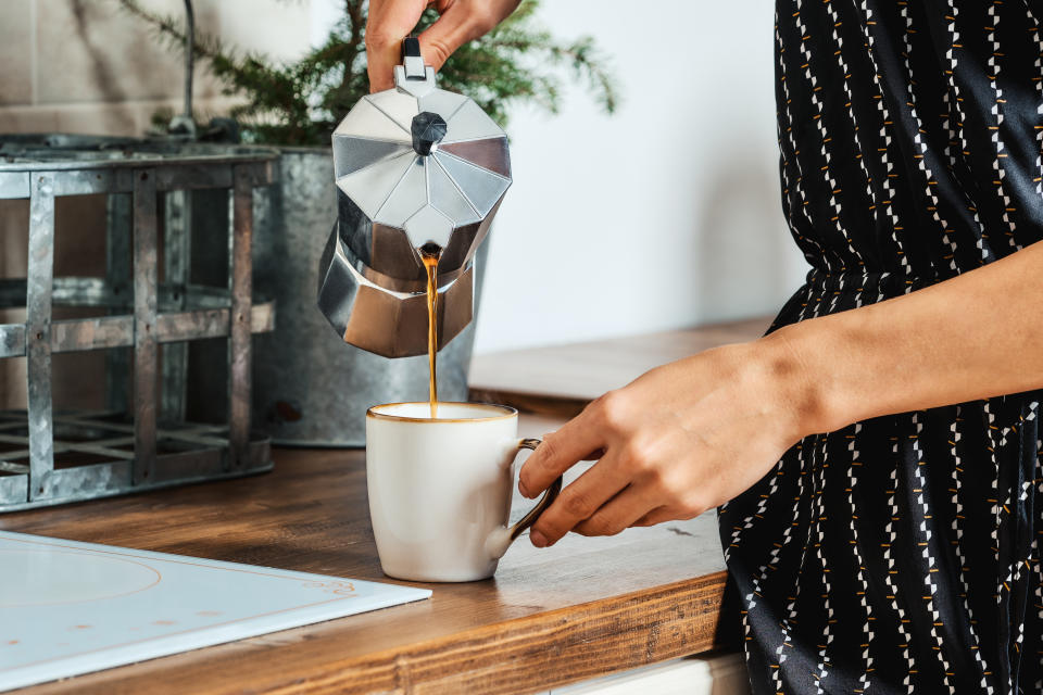 A person in patterned clothing is pouring coffee into a white mug from a moka pot on a kitchen counter with a plant in the background