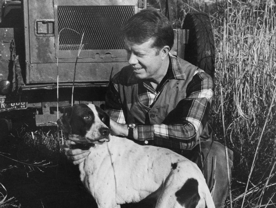 Jimmy Carter crouching down to pet his dog behind an unspecified tractor on his peanut farm in Plains, Georgia, 1970