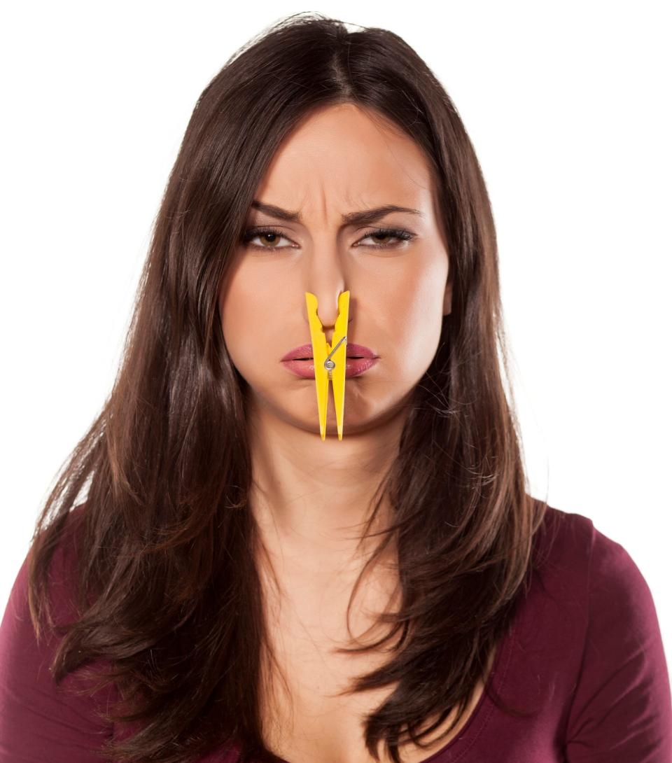 A woman with long hair has a yellow clothespin clipped on her nose with a displeased expression