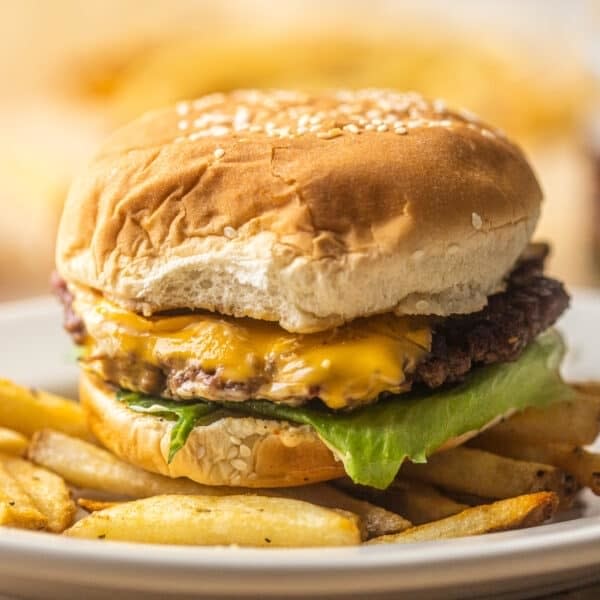 A cheeseburger with lettuce on a sesame seed bun, served on a white plate with a side of French fries