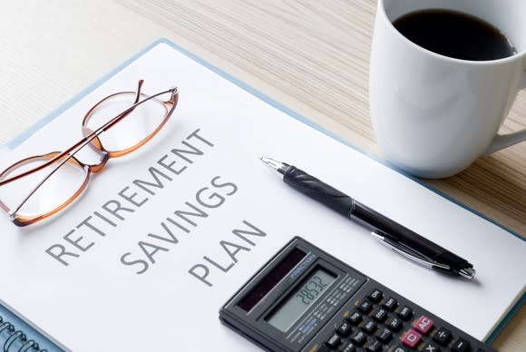 Binder labeled "RETIREMENT SAVINGS PLAN" on a table with a pair of glasses, a calculator, and a pen on top of it and a coffee cup standing next to it. 