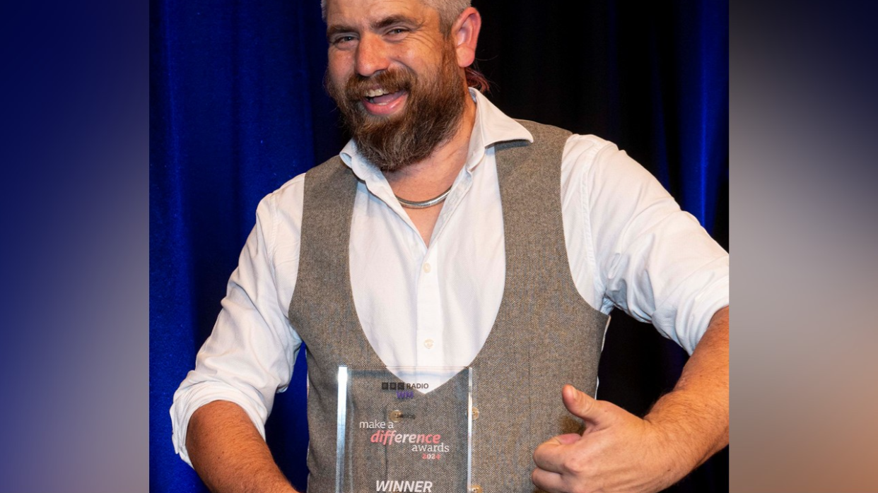 Ben Newman, with bear and brown hair, is wearing a white shirt and brown waistcoat and is smiling and giving the thumbs up while holding his award