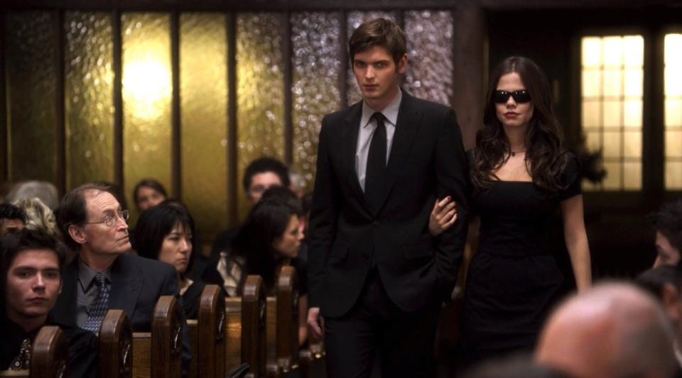 A man in a suit is walking down a church aisle with Jenna, who is dressed in black and wearing dark sunglasses