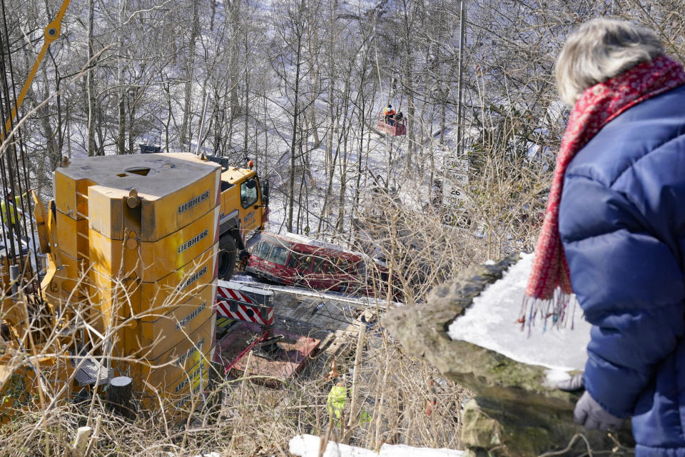 A passerby, right, watches as workers in a basket suspended by a crane help with the recovery process on Monday Jan. 31, 2022 of the bus and other vehicles that were on a bridge when it collapsed Friday, in Pittsburgh's East End. (AP Photo/Gene J. Puskar)