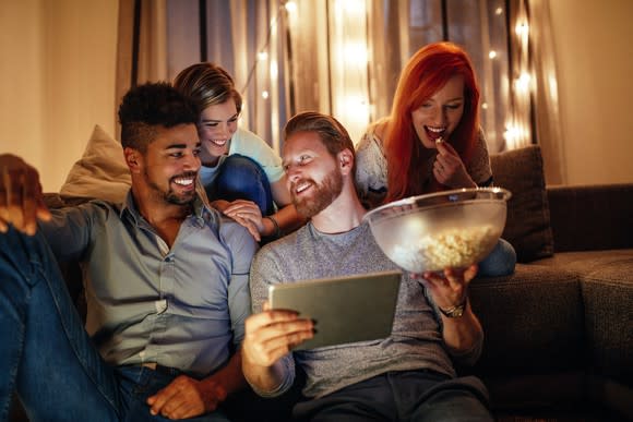 Friends sitting on a couch and on the floor. One holds a tablet and a bowl of popcorn.