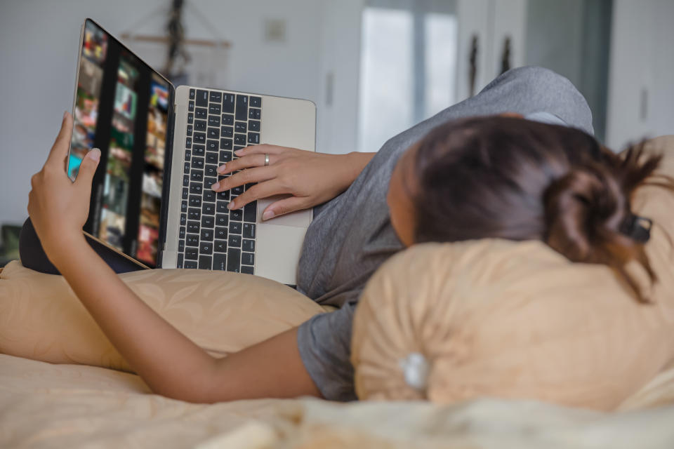 Woman holding laptop lying on bed looking at TV shows