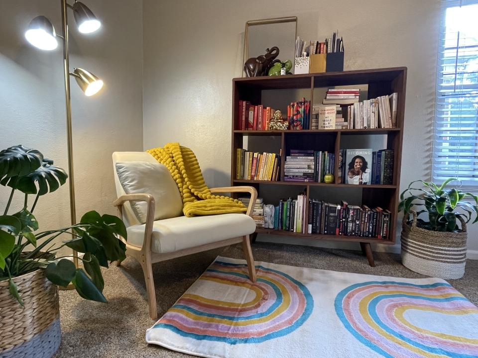 Comfortable reading nook with a cozy armchair, a yellow throw blanket, a colorful rug, bookshelves filled with various books, and several potted plants