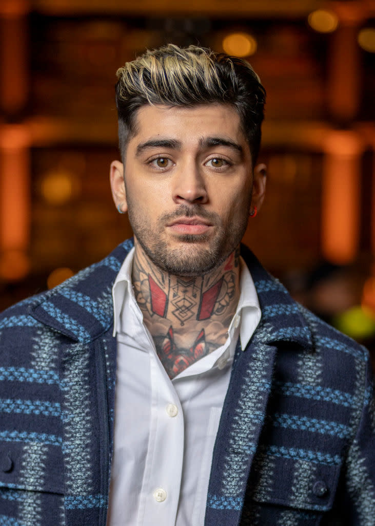 Zayn Malik, dressed in a white shirt and a jacket with a checkered pattern, stands with a serious expression. Tattoos are visible on his neck