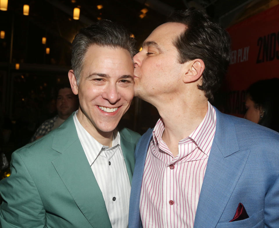 Jim Parsons kisses his partner Todd Spiewak on the cheek, both smiling at a public event. Jim wears a blue blazer, and Todd is in a green suit