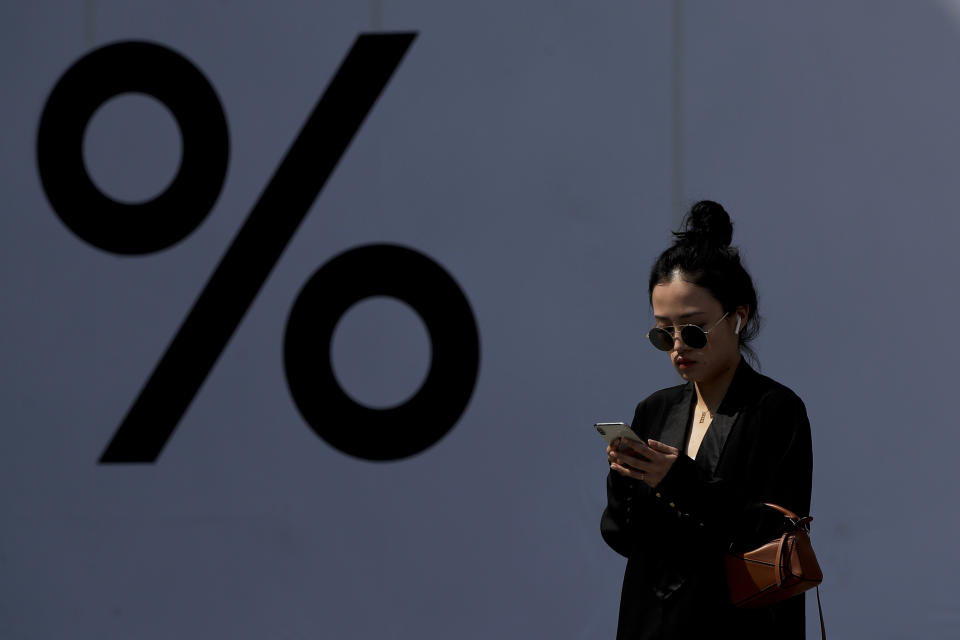 A Chinese woman wearing an Apple AirPods while browsing her iPhone past by a booth bearing a percent sign at the capital city's popular shopping mall in Beijing, Thursday, April 4, 2019. The U.S. and China opened a ninth round of talks on Wednesday, aiming to further narrow differences in an ongoing trade war that has deepened uncertainty for businesses and investors and cast a pall over the outlook for the global economy. (AP Photo/Andy Wong)