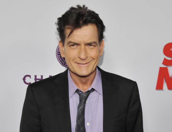 Charlie Sheen smiles and poses while wearing a dark suit and light purple shirt with a gray patterned tie