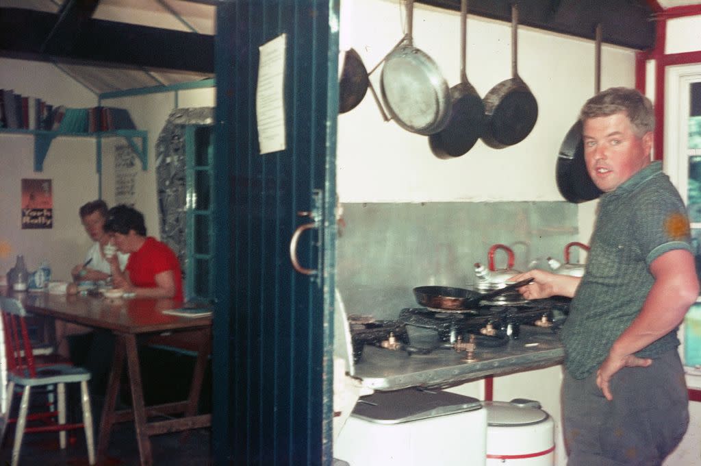 Vernacular photograph taken on a 35mm analog film transparency, believed to depict black and silver frying pan on stove, with man in the processing of cooking, in the kitchen of a restaurant or hostel, 1970