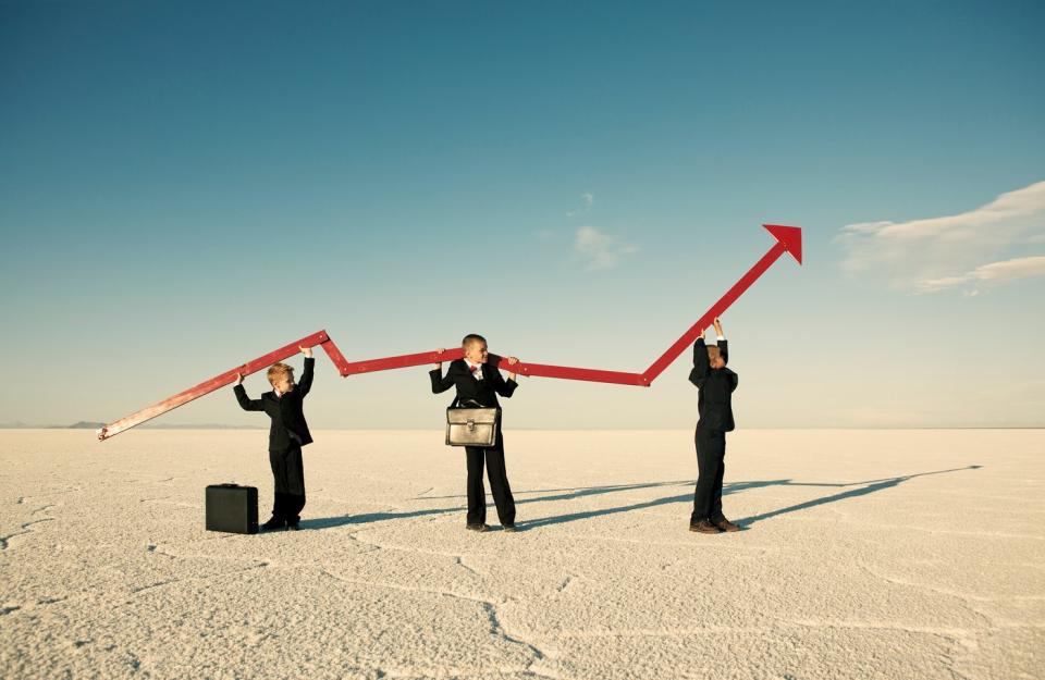 Three kids dressed as businessmen raise a rising chart in the middle of a desert.