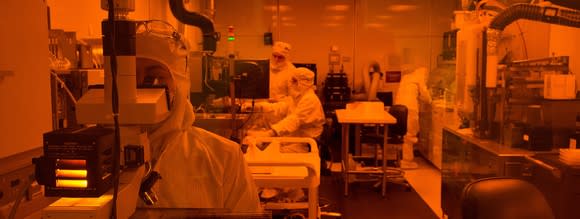 Lab in orange light with people wearing masks and sterile clothing working on various types of equipment.