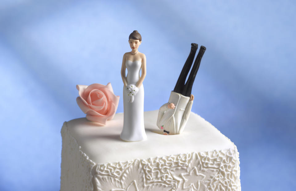 A bride in a white dress stands next to an upside-down groom figurine on a wedding cake decorated with a single rose