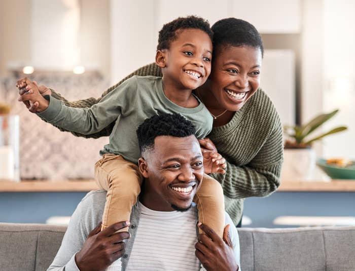 A smiling family of three, with a child sitting on an adult's shoulders, posing happily in a cozy indoor setting