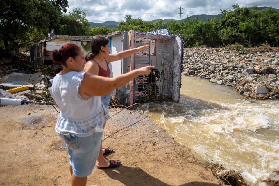 Two women point toward a river next to a house destroyed by flooding.