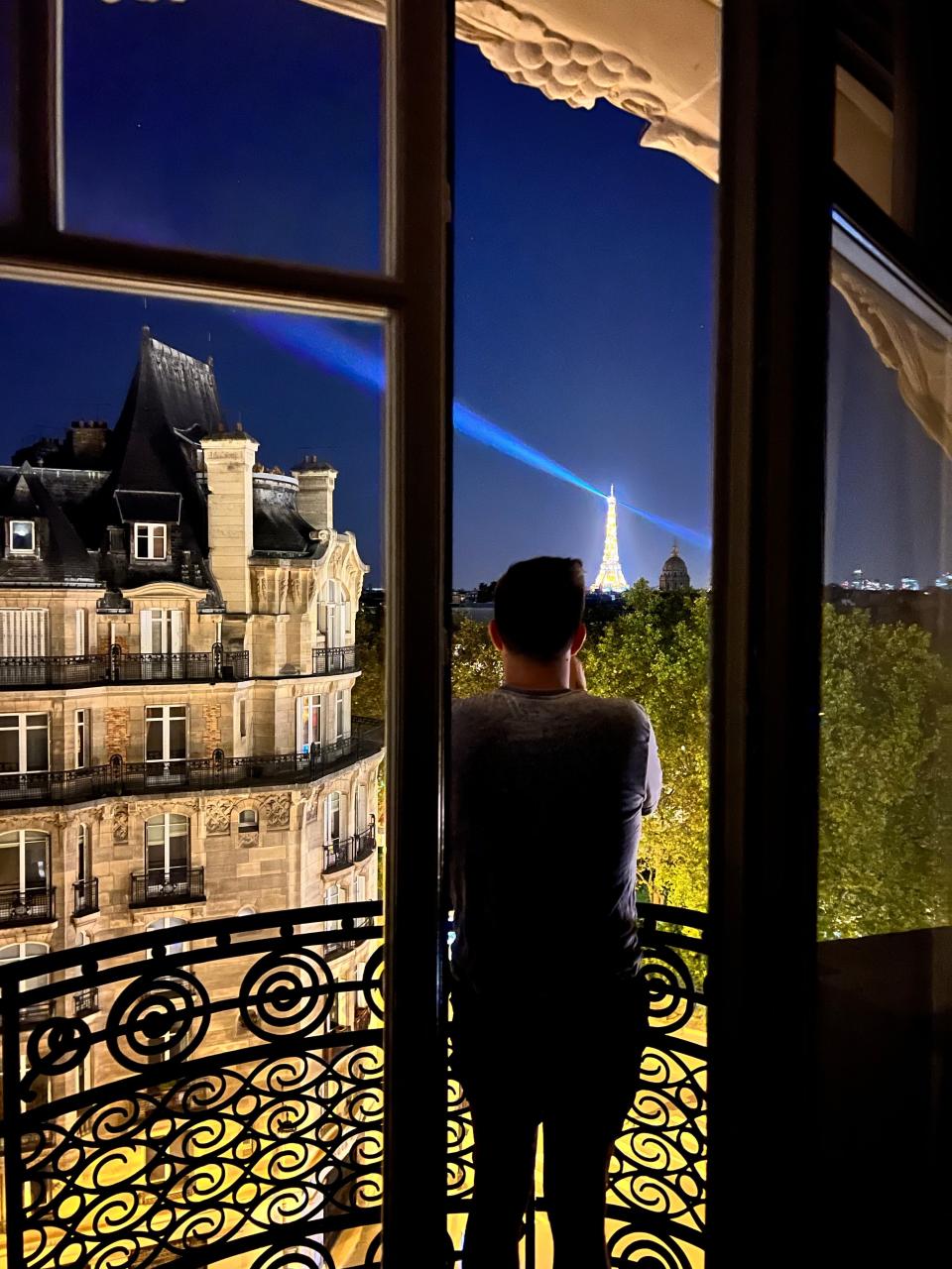 A man standing on a balcony at night taking a picture of the Eiffel Tower.