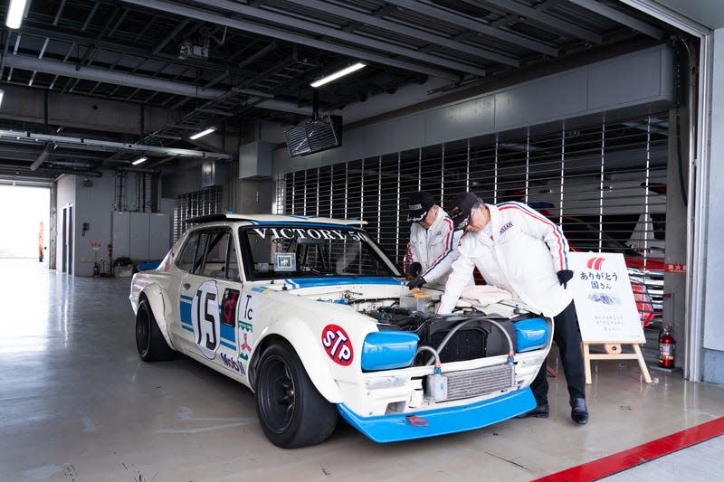 a white and blue 1972 gt-r in a garage, being worked on by two older men in clothing from the car's period