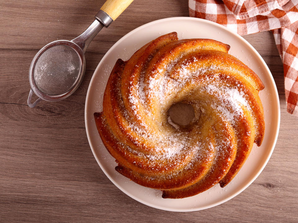 A circular Bundt cake with powdered sugar on top, placed on a wooden table next to a fine mesh strainer and a checkered cloth