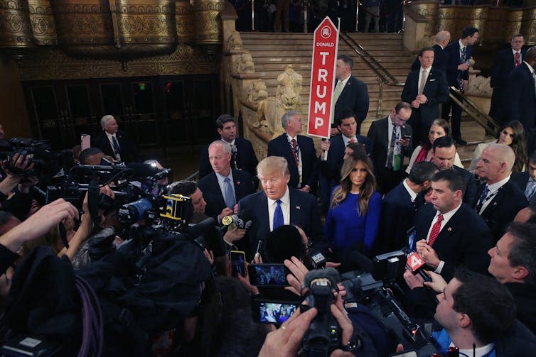 A man in a white shirt, dark jacket and tie talking to a crush of reporters.