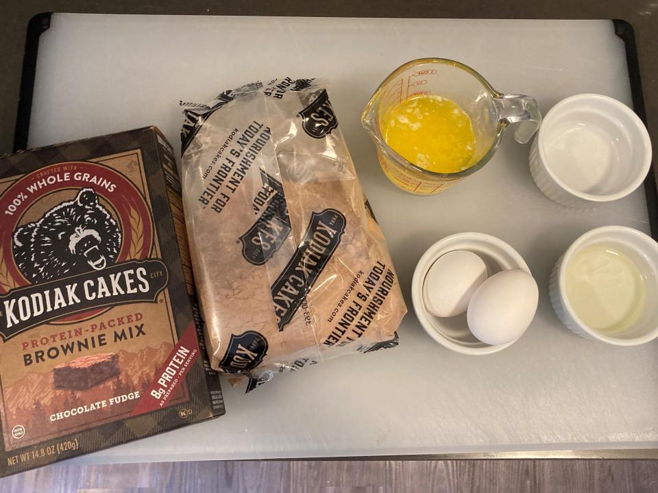 Kodiak Cakes brownie mix box, package of brownie mix, liquid measuring cup with melted butter, cup of water, cup of oil, and bowl with two eggs on cutting board