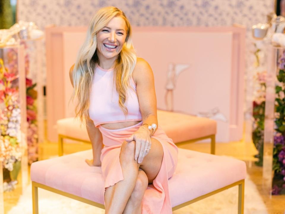 Hayley Paige sits on a pink ottoman wearing fluffy shoes.