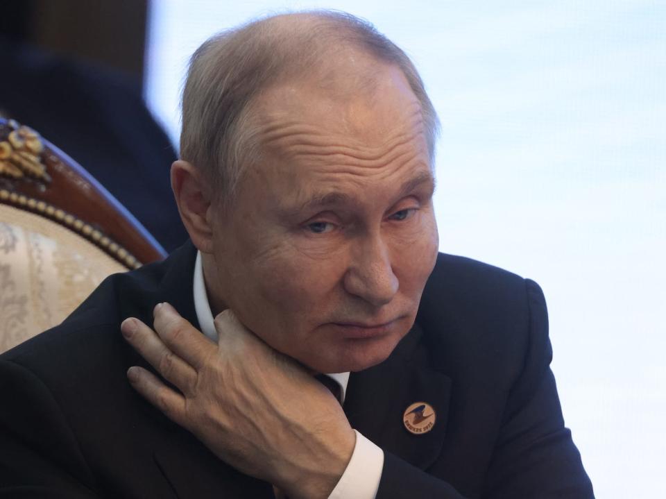 Russian President Vladimir Putin adjusting the collar of his shirt while sitting in a wooden chair with gold accents.